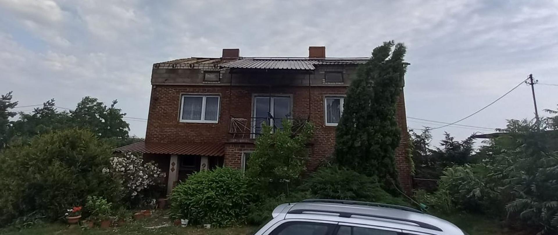 Na zdjęciu widać uszkodzony dach budynku mieszkalnego z czerwonej cegły. Wokół domu widać drzewa i krzewy. Przed domem widać dach zaparkowanego srebrnego samochodu osobowego. Jest dzień.