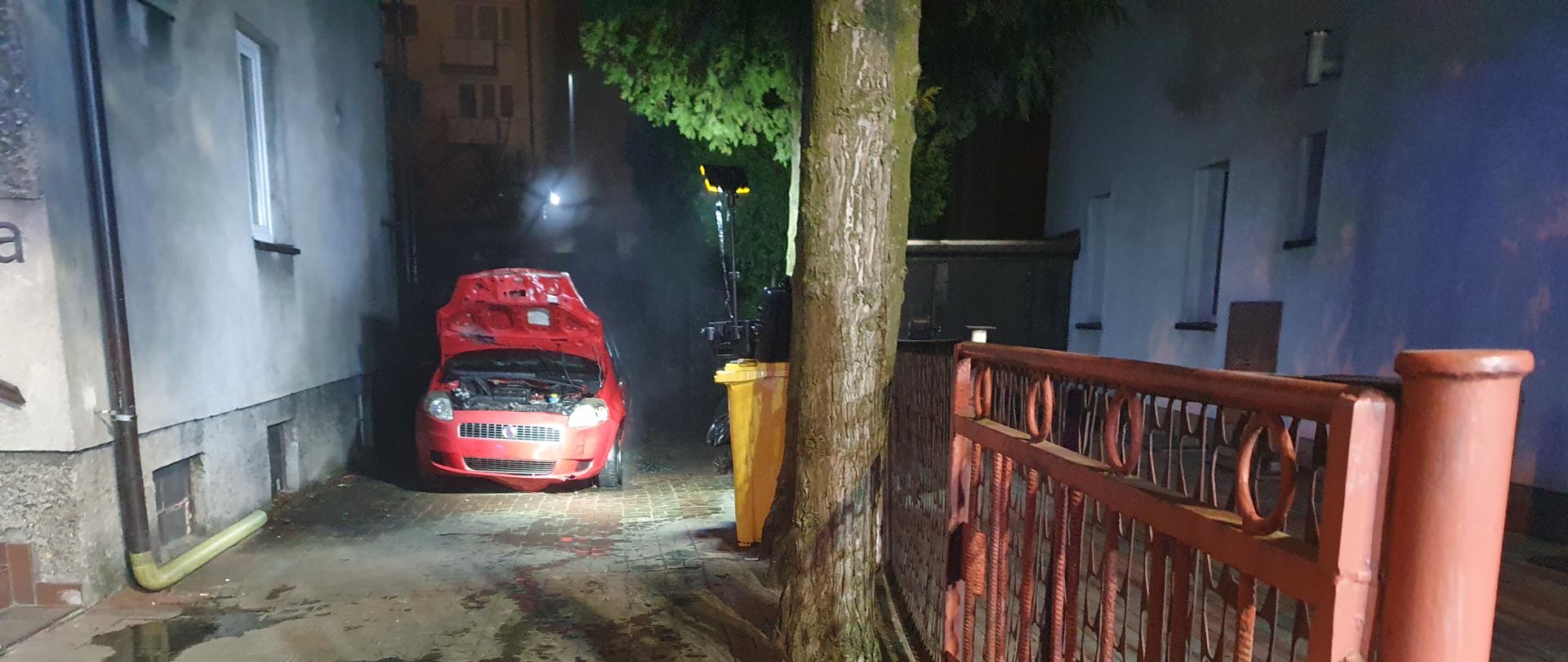 Spalony wrak samochodu. Widok z przodu. Samochód ma otwartą maskę. Jest zaparkowany na posesji. Z lewej strony budynek mieszkalny. 