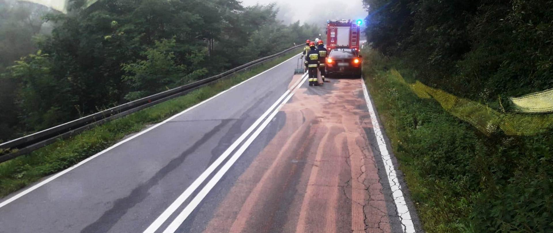 Samochód pożarniczy na pasie drogi a przed nim i smuga z sorbentu.Obok samochodu strażacy.