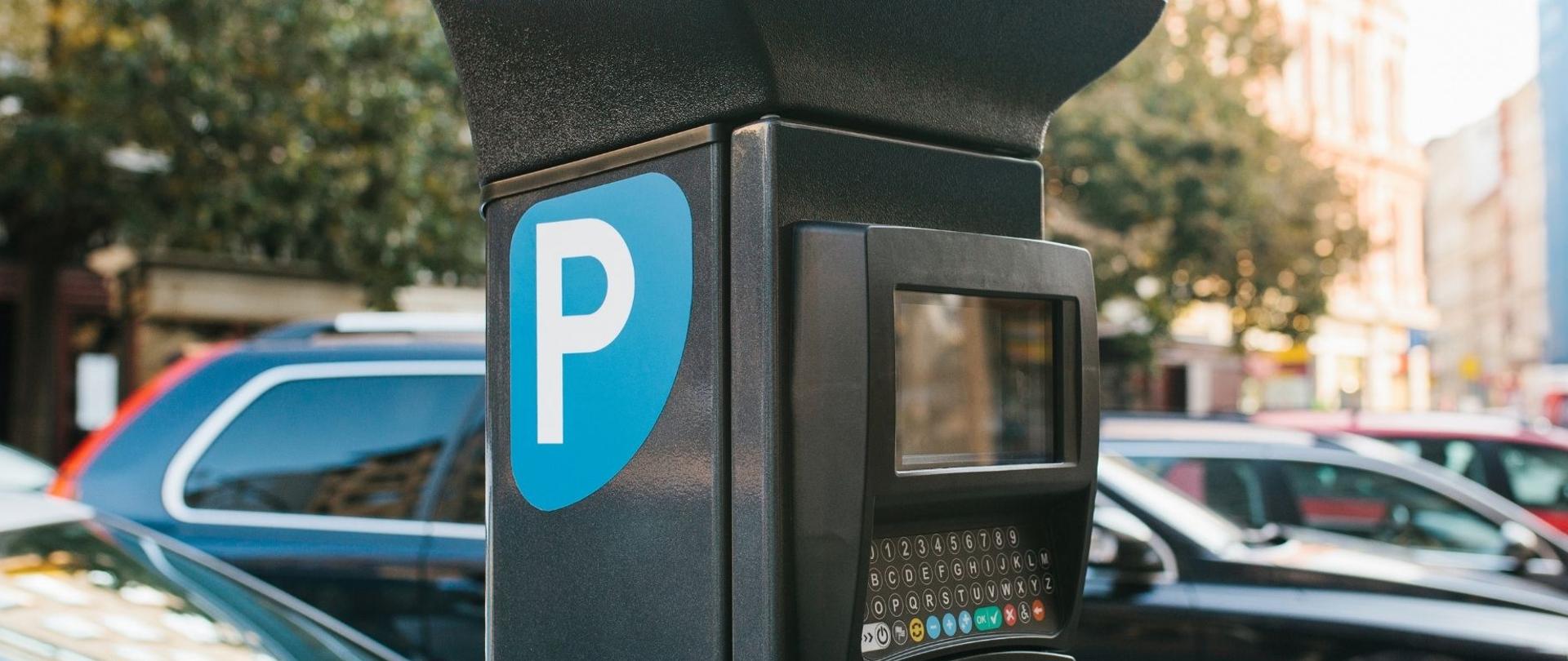 Zdjęcie przedstawia: na pierwszym planie fragment parkometru znajdującego się w strefie płatnego parkowania niestrzeżonego w Warszawie. W tle znajduje się szereg zaparkowanych samochodów.