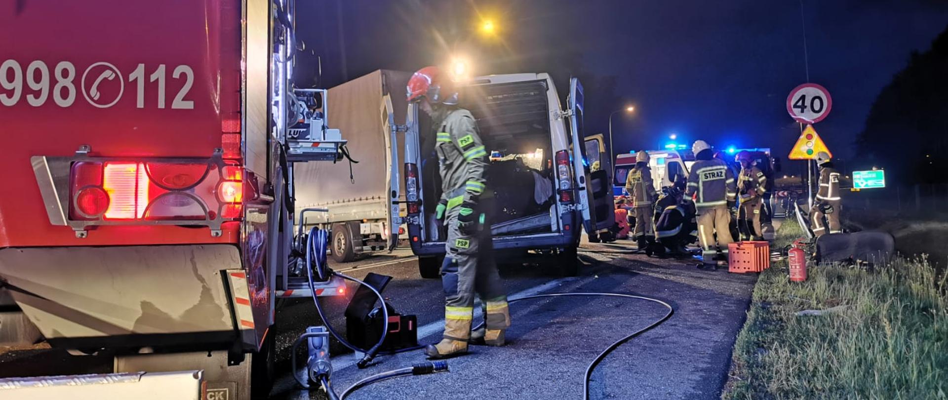 Widok stojących na jezdni pojazdu pożarniczego, dwóch busów jeden za drugim oraz umundurowanych strażaków.
