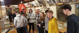 Młodzież stoi w Sali pełnej fotografii i pamiątek. Kustosz muzeum pokazuje poszczególne eksponaty z czasów II wojny światowej, broń mundury, rzeczy osobiste, dokumenty.