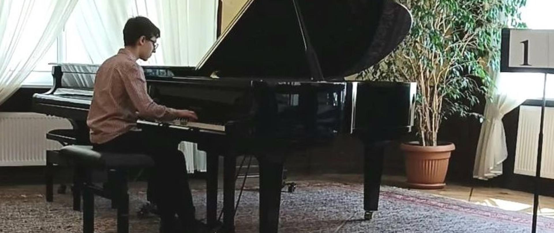 Zdjęcie kolorowe. Chłopak o ciemnych włosach, w okularach, ubrany w koszulę i czarne spodnie gra na czarnym fortepianie podczas występu na konkursie pianistycznym. Po prawej stronie fortepianu stoi duży kwiat doniczkowy. W tle okna ozdobione białą firaną i beżową zasłoną.