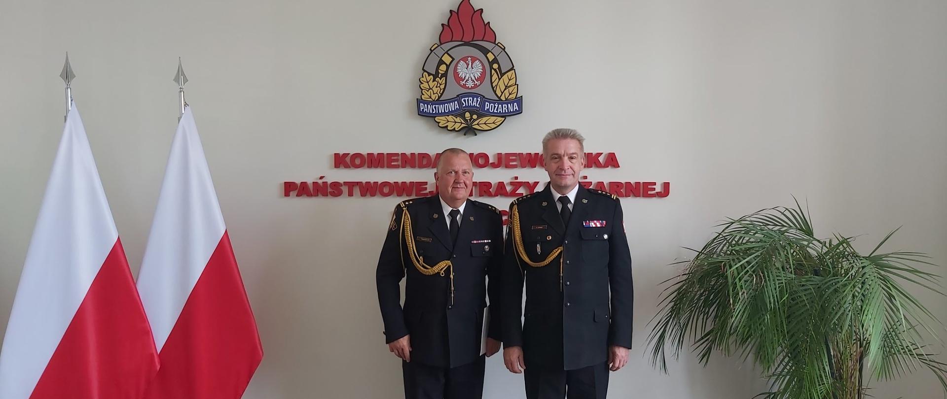 Po prawej st.bryg. Kazimierz Lesisz w mundurze wyjściowym, po lewej bryg. Jacek Twardoń w mundurze wyjściowym. W tle znajduje się ściana pamiątkowa z napisem "Komenda Wojewódzka Państwowej Straży Pożarnej w Szczecinie" wraz z logo PSP, po lewej znajdują się flagi, po prawej kwiat.