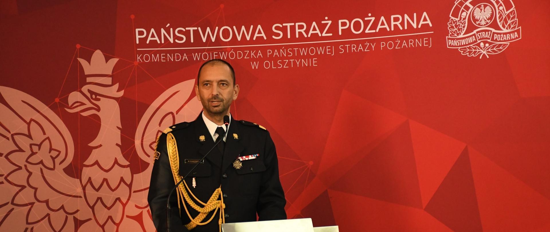 Na zdjęciu warmińsko mazurski komendant wojewódzki w mundurze galowym, stoi przy mównicy. W tle czerwona ścianka z napisem Państwowa straż pożarna.