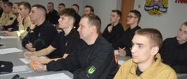 Świetlica KP PSP w Rawiczu. Za rzędami stołów zasiadają słuchacze - uczestnicy szkolenia podstawowego OSP. Mają na sobie koszulki w kolorze czarnym, ubrania koszarowe w kolorze czarnym lub piaskowym. Wśród słuchaczy są mężczyźni. Rozwiązują test pisemny, mając przed sobą arkusze egzaminacyjne. W tle ściana z logo PSP.
