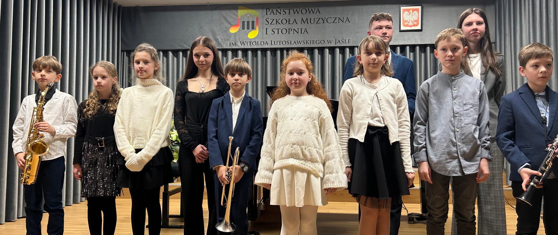 Grupa uczniów Państwowej Szkoły Muzycznej I stopnia im. Witolda Lutosławskiego w Jaśle stoi na scenie podczas szkolnego koncertu. Dzieci ubrane są w stroje galowe; część z nich trzyma instrumenty dęte – saksofon, trąbkę i klarnet. W tle widoczna jest kurtyna oraz tablica z nazwą szkoły i godłem Polski.