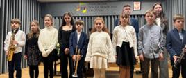 Grupa uczniów Państwowej Szkoły Muzycznej I stopnia im. Witolda Lutosławskiego w Jaśle stoi na scenie podczas szkolnego koncertu. Dzieci ubrane są w stroje galowe; część z nich trzyma instrumenty dęte – saksofon, trąbkę i klarnet. W tle widoczna jest kurtyna oraz tablica z nazwą szkoły i godłem Polski.