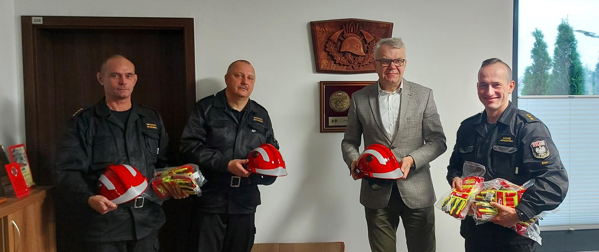 Zdjęcie przedstawia dyrektora firmy Novago przekazującego środki ochrony indywidualnej dla żnińskich strażaków w postaci rękawic technicznych oraz hełmów Komendantowi Powiatowemu i Dowódcom Jednostki Ratowniczo-Gaśniczej