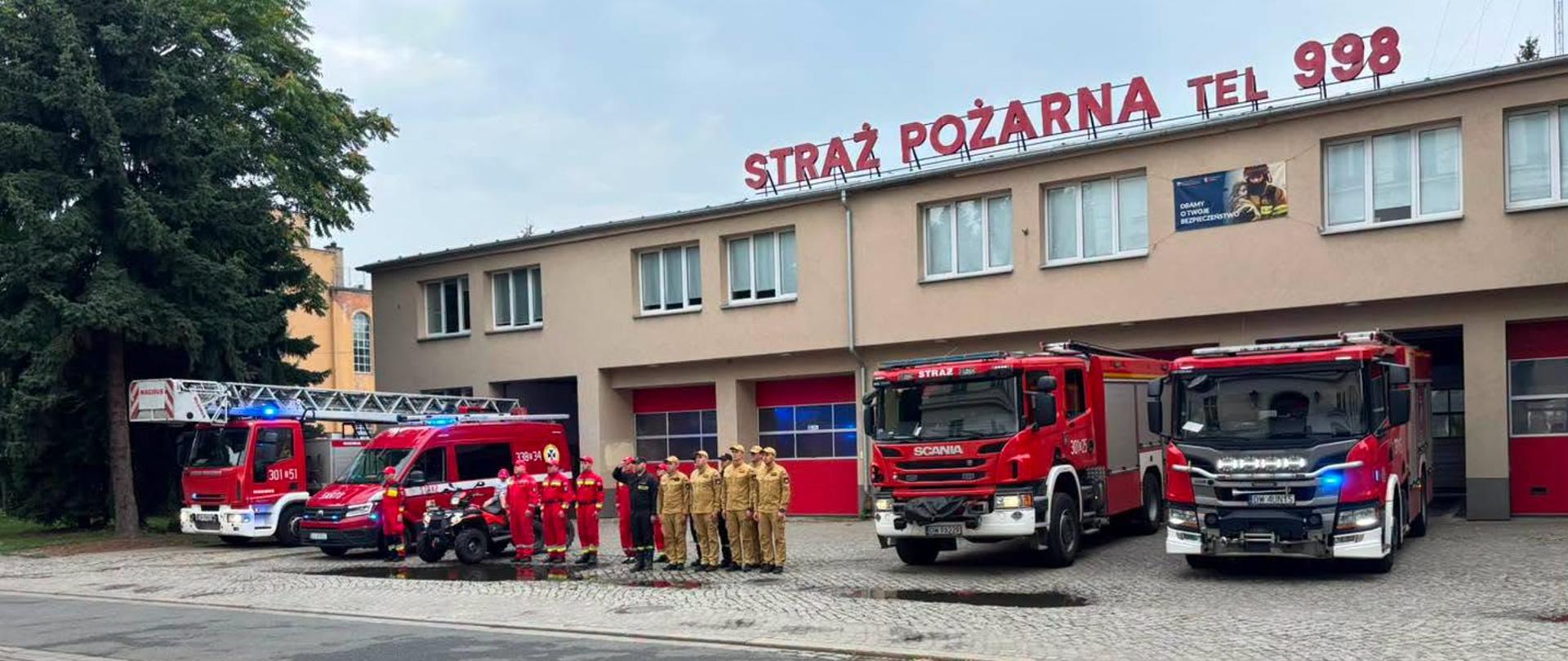 Zdjęcie przedstawia remizę strażacką z napisem „STRAŻ POŻARNA TEL 998” na dachu. Przed budynkiem zaparkowane są trzy pojazdy straży pożarnej: stary, zabytkowy wóz strażacki po lewej i dwa nowoczesne wozu strażackie po prawej. Grupa strażaków stoi przed zabytkowym wozem. Na pierwszym planie widać tory tramwajowe.