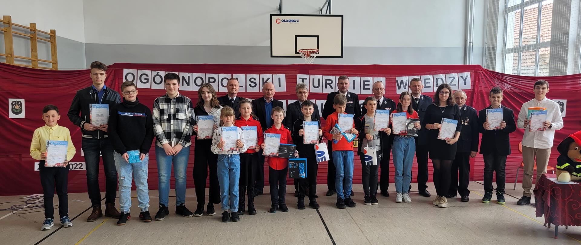 grupa dzieci trzymająca dyplomy w sali gimnastycznej stojąca w szeregu wraz ze strażakami, w tle widoczny napis : ogólnopolski turniej wiedzy pożarniczej