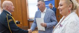 Mężczyzna w szarym garniturze trzyma dokument i wita się uściskiem dłoni ze strażakiem w mundurze galowym Państwowej Straży Pożarnej. Obok nich stoi uśmiechnięta kobieta w białej, koronkowej marynarce, która przygląda się powitaniu