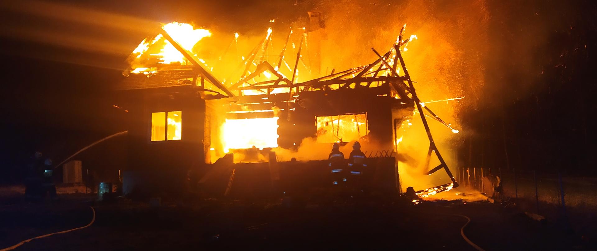 Zdjęcie przedstawia budynek objęty w całości ogniem. Wokół domu widnieje żółta poświata. Na tle płomieni można zauważyć resztki więźby dachowej. Widać również strażaków gaszących pożar.
