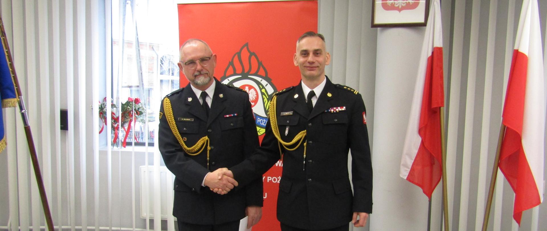 Zdjęcie zrobione w pomieszczeniu strażnicy na tle na tle godła Rzeczpospolitej Polskiej, Sztandaru Komendy Powiatowej Państwowej Straży Pożarnej w Lublińcu, dwóch Flag Rzeczpospolitej Polskiej oraz baneru Komendy Powiatowej PSP w Lublińcu. Na pierwszym planie Komendant Powiatowy Państwowej Straży Pożarnej w Lublińcu st. bryg. Janusz Bula z uściskiem dłoni z funkcjonariuszem odchodzącym na zaopatrzenie emerytalne. Strażacy w umundurowaniu galowym.