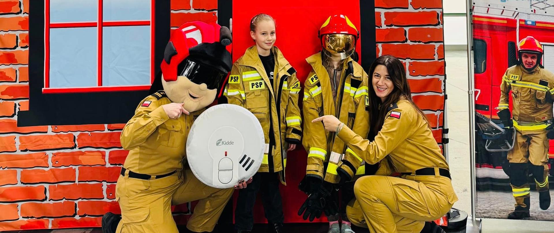 maskotka strażak Ejber z czujką i kobieta strażak wskazują na czujkę, między nimi stoi dwójka dzieci w hełmie i mundurach 