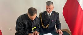 Nowoprzyjęty strazak podpisuje akt ślubowania obok siedzi Komendant Powiatowy
