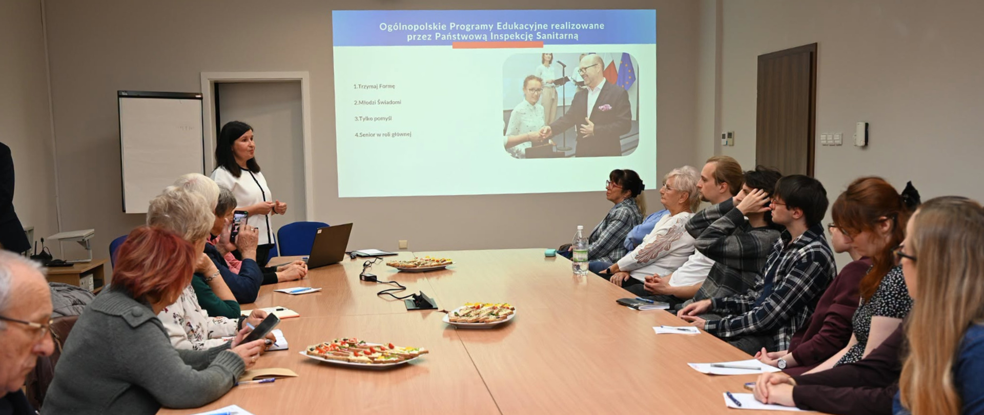 Agnieszka Noculak z WSSE w Bydgoszczy prowadzi prezentację dotyczącą programów edukacyjnych Państwowej Inspekcji Sanitarnej. Uczestnicy spotkania, wśród których są seniorzy i młodzież, siedzą przy wspólnym stole i słuchają wystąpienia. W tle wyświetlana jest prezentacja multimedialna poświęcona profilaktyce zdrowotnej.