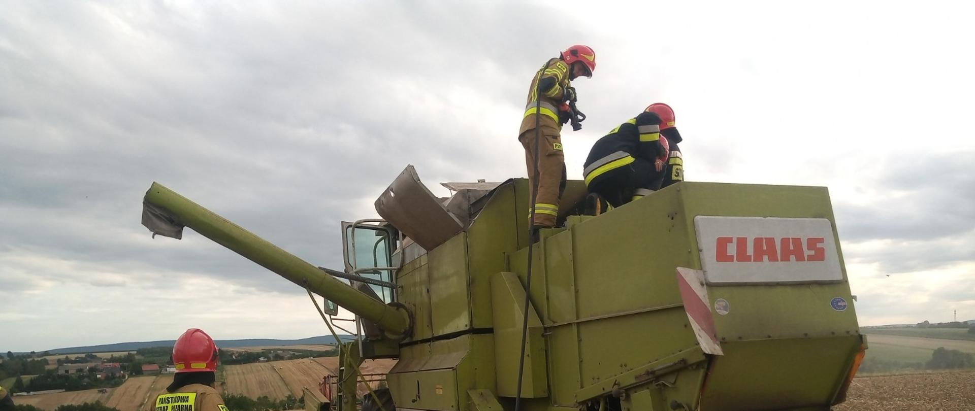 Kombajn zbożowy na polu uprawnym, na dachu pojazdu dwóch strażaków dogaszających komorę silnika
