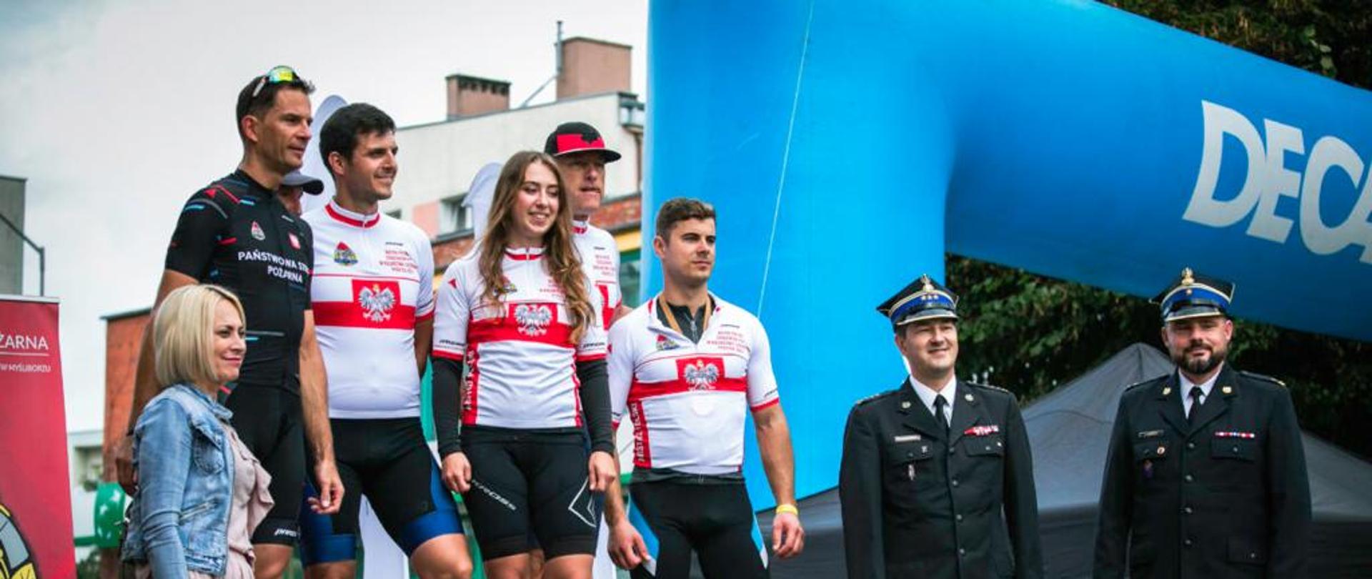 Na podium 5 zawodników w tym 1 kobieta w czarnych getrach i biało czerwonych koszulkach z znakiem orła, po prawej stronie stoi dwóch funkcjonariuszy PSP, po lewo kobieta 
