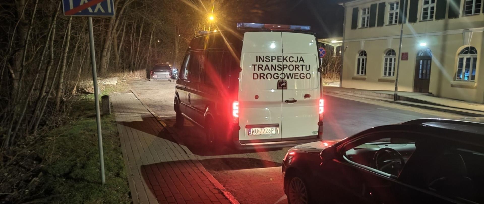 Nocna scena uliczna oświetlona latarniami. Na poboczu stoi pojazd z napisem Inspekcja Transportu Drogowego, z włączonymi światłami. Za nim jest ciemny samochód osobowy. Obok znajduje się chodnik z kostki brukowej oraz znak z przekreślonym napisem TAXI. W tle widoczne drzewa bez liści oraz zaparkowany pojazdy i budynek.