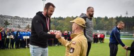 Mężczyzna w czarnej bluzie i jeansach, ze medalem na szyi, ściska dłoń mężczyzny w beżowym mundurze na podium, podczas ceremonii wręczenia nagród.