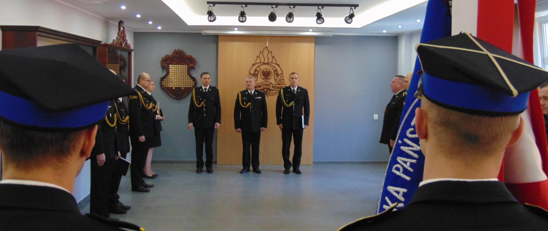 Świetlica komendy. Po prawej stronie strażacy stojacy w dwuszeregu. Po środku komendant ora odchodzący i przejmujący dowódca. po lewej oficerowie prowadzący zbiórkę. Tyłem do fotografa poczet ze sztandarem