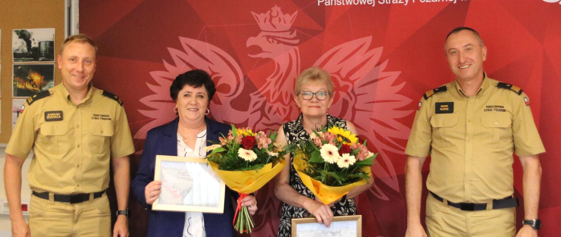  Na zdjęciu widoczne są cztery osoby. W centrum stoją dwie pracownice w schludnych ubraniach cywilnych, które odchodzą na emeryturę. Po obu ich stronach znajdują się dwaj funkcjonariusze w piaskowych mundurach służbowych. Kobiety trzymają w rękach kwiaty oraz pamiątkowe dyplomy w drewnianych ramkach. Wszyscy uśmiechają się i patrzą w stronę fotografa. W tle widoczna jest czerwona ikonografia Państwowej Straży Pożarnej wraz z emblematem Szkoły Aspirantów PSP w Krakowie.