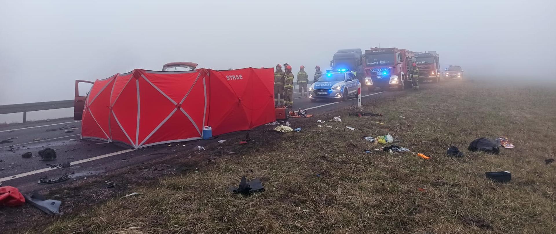 W centralnej części fotografii, widać stojący na drodze czerwony strażacki parawan, zasłaniający rozbite auto. Z prawej strony widoczne sylwetki strażaków, a za nimi pojazdy służb ratowniczych. Do około porozrzucane elementy karoserii samochodów.