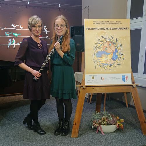 na zdjęciu widoczna jest uczennica i jej nauczycielka trzymające klarnet, po prawej stronie znajduje się plakat o treści Festiwal Muzyki Słowiańskiej