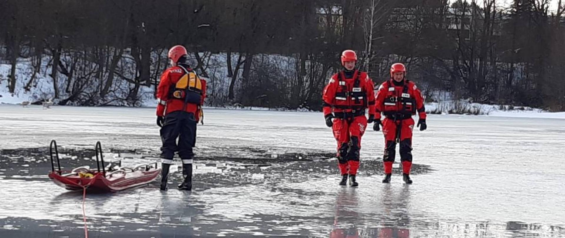 Zdjęcie przedstawia 3 strażaków ubranych w skafandry nurkowe oraz kaski. Strażacy poruszają się po zamarzniętym zbiorniku wodnym - chodzą po lodzie. Do ćwiczeń wykorzystują sanie lodowe.