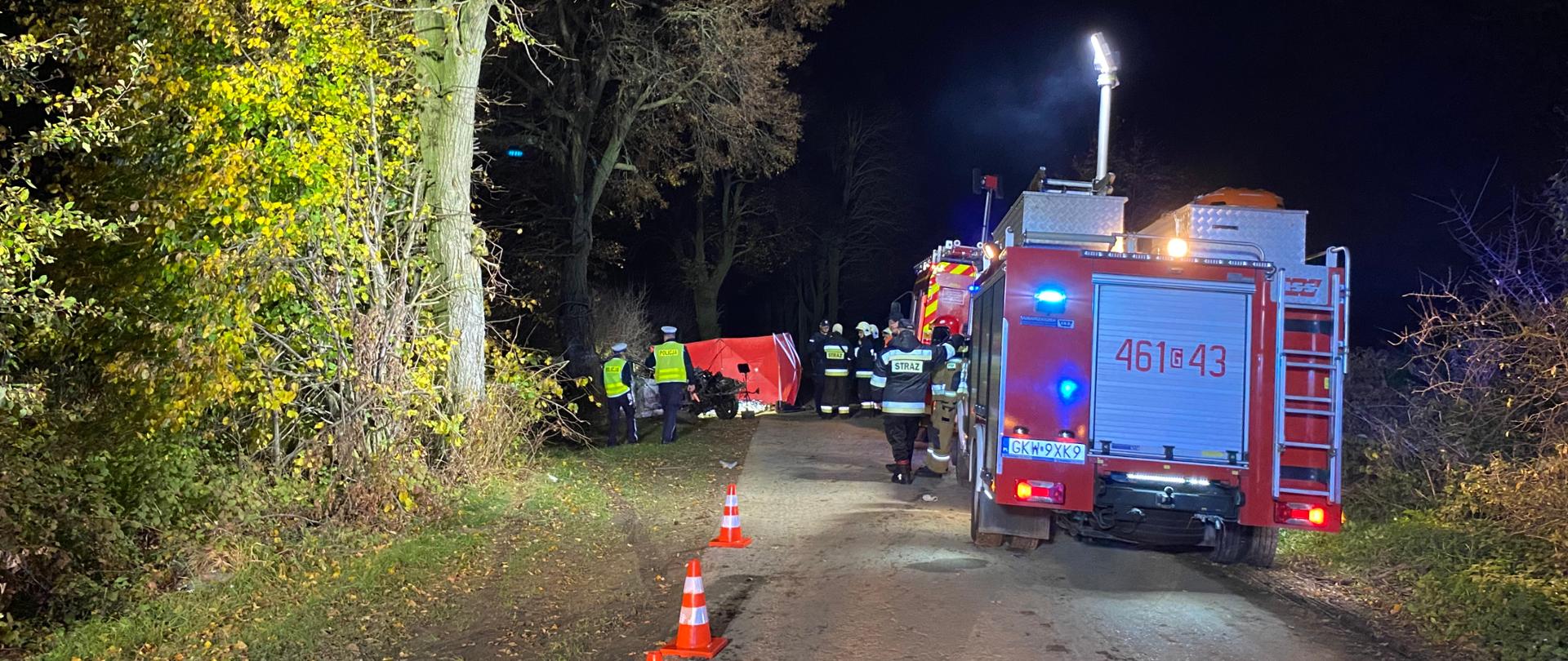 Samochody ratownicze stoją na jezdni w porze nocnej w oddali parawan i wrak pojazdu