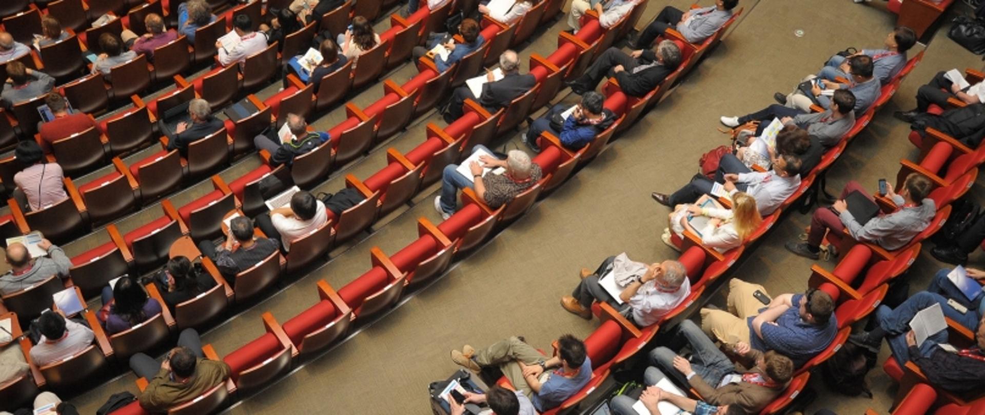 Sala konferencyjna z uczestnikami siedzącymi w czerwonych krzesłach, uczestniczącymi w wykładzie lub spotkaniu.