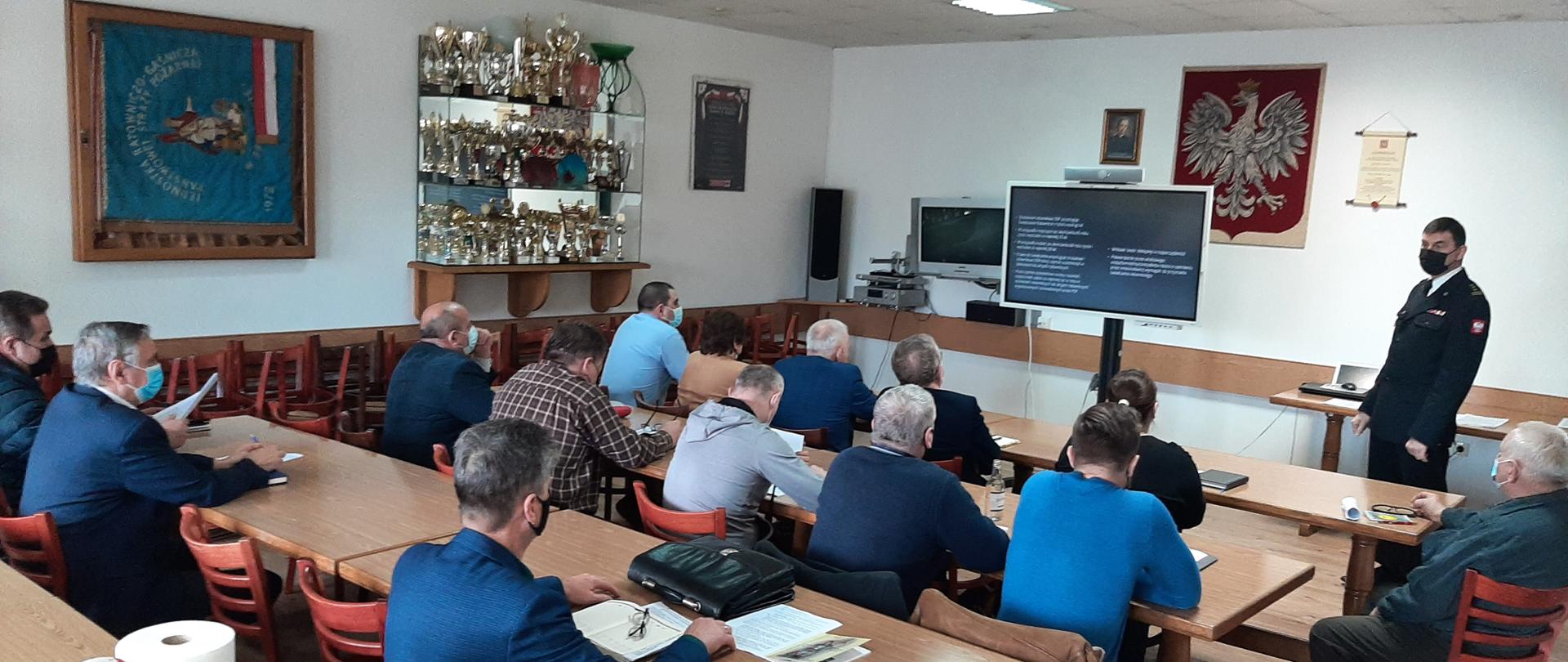 Kolorowa fotografia przedstawia wnętrze dużej sali w budynku. Na pierwszym planie kilkanaście osób cywilnych siedzi przy stołach, w maskach i obserwuje prezentację. Po prawej stronie stoi umundurowany na wyjściowo strażak w masce, który prowadzi prezentację. Na środku sali duży monitor wyświetlający treść prezentacji. Na ścianach jasnej sali liczne puchary, sztandar, godło i inne obrazy.
