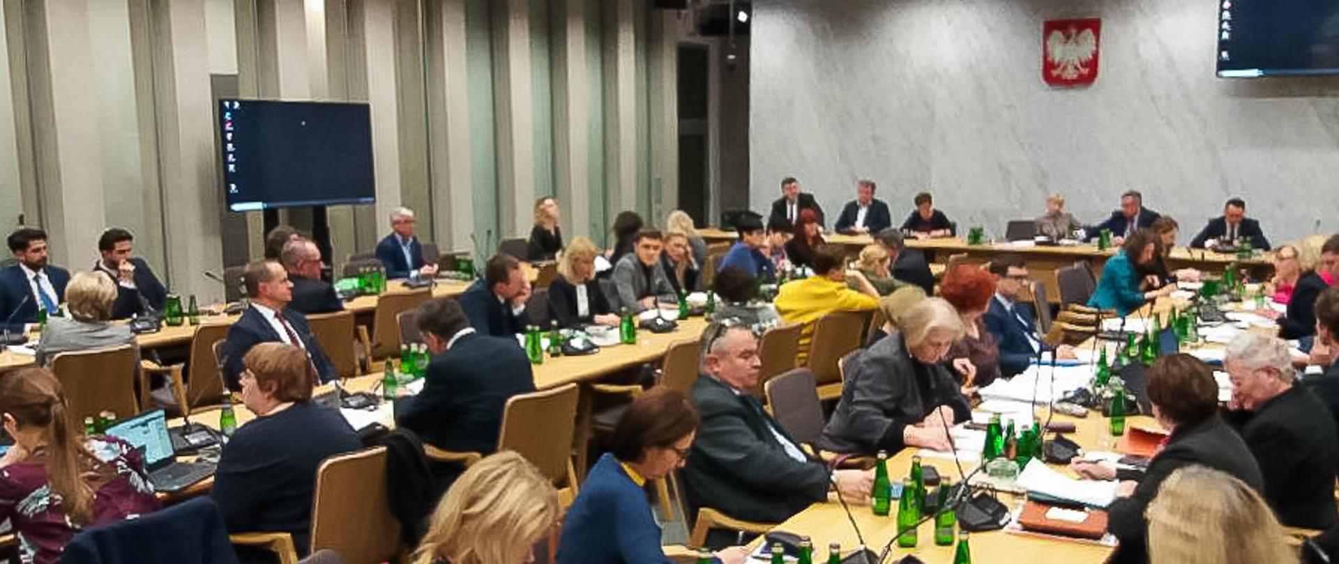 Posiedzenie Sejmowej Komisji Edukacji, Nauki i Młodzieży z udziałem ministra edukacji. Ludzie siedzą przy stołach w dużej sali i dyskutują.