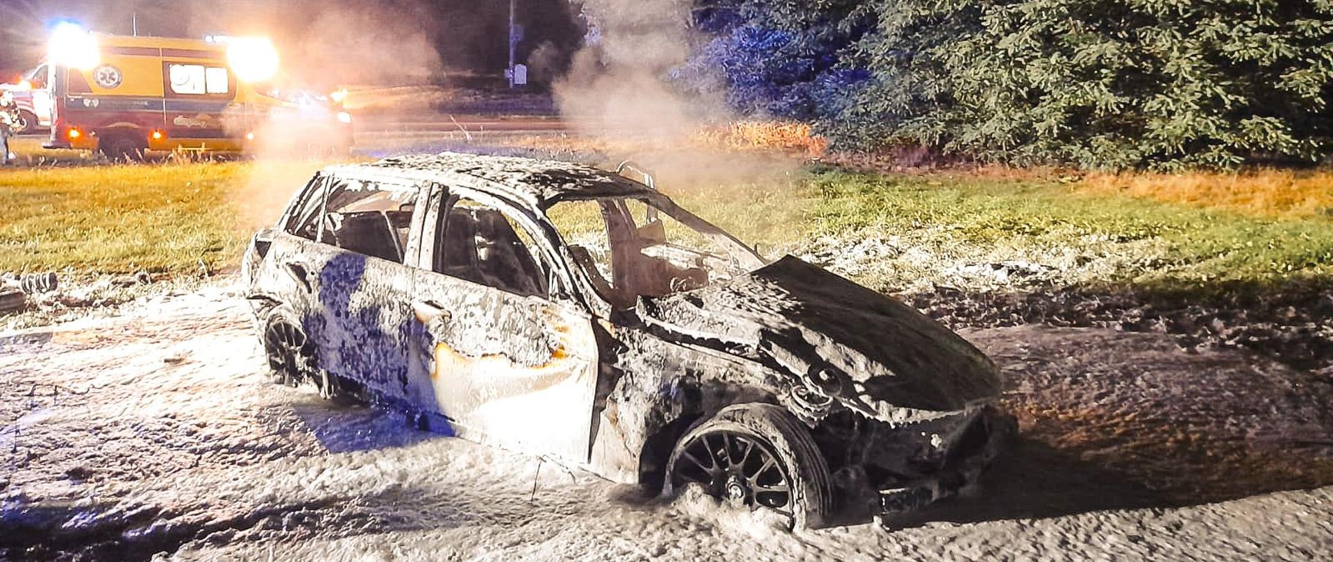 Całkowicie spalony samochód osobowy znajduje się na pasie zieleni. Na drugim planie widać ambulans Ratownictwa Medycznego oraz po prawej stronie las. Zdjęcie wykonane w porze nocnej.