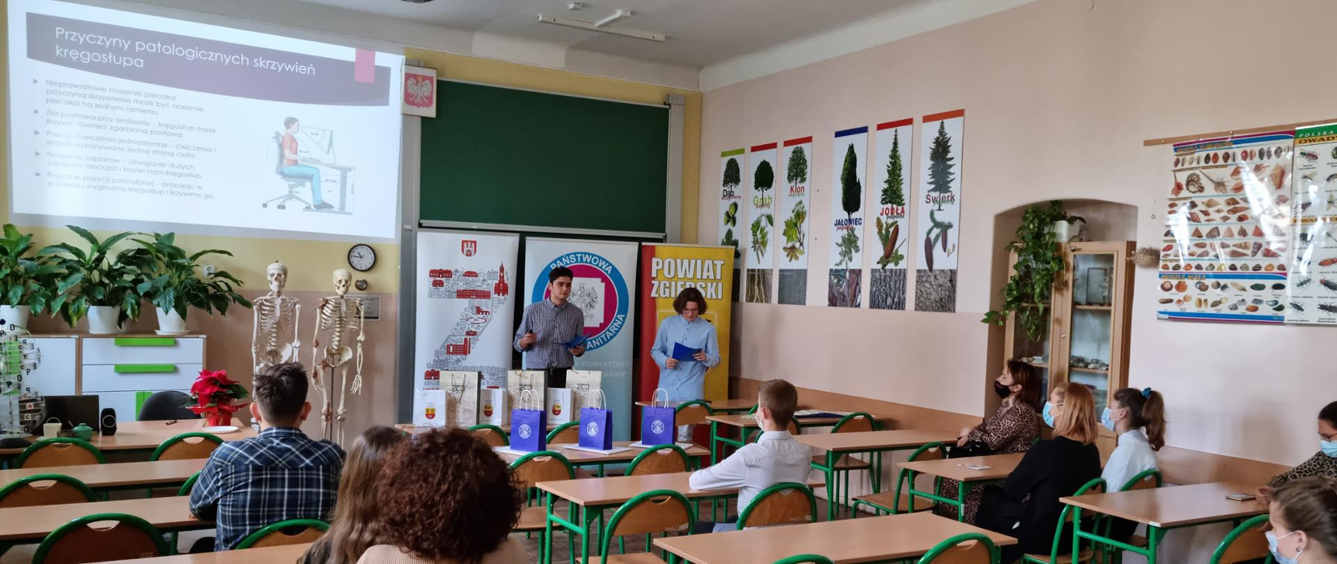 Zdjęcie zrobione jest w szkole w pracowni biologicznej. W sali znajdują się dorośli i młodzież. Dwóch uczniów prowadzi wykład. Na tablicy wyświetla się slajd o przyczynach patologicznych skrzywień kręgosłupa