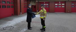 Zastępca Komendanta Powiatowego PSP w Rypinie starszy brygadier Andrzej Górecki wręcza list gratulacyjny starszemu ogniomistrzowi Krystianowi Sowińskiemu z okazji dwudziestopięciolecia służby i pracy. W tle i po lewej stronie zdjęcia czerwone bramy garaże.