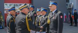 
Na zdjęciu moment wręczenie odznaczenia strażakom przez dwóch innych strażaków w mundurach wyjściowych koloru czarnego na tle budynku.