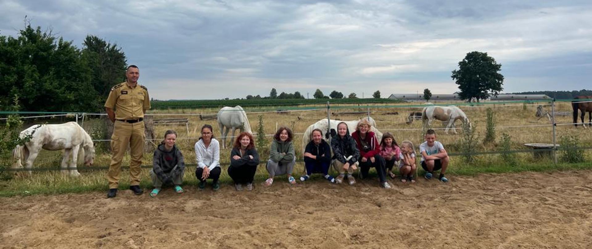 dzieci ze strażakiem podczas zbiórki w tle las i pasące się konie 