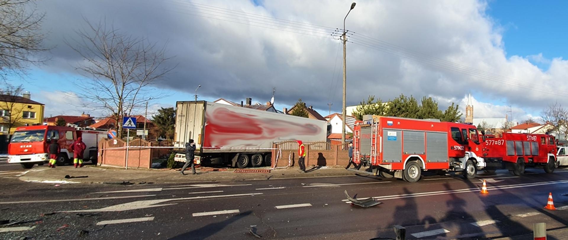 Ulica na której rozrzucone są elementy powypadkowe. Na drugim planie ciężarówka, która wjechała na posesję, po lewe i prawej stronie samochody pożarnicze. 