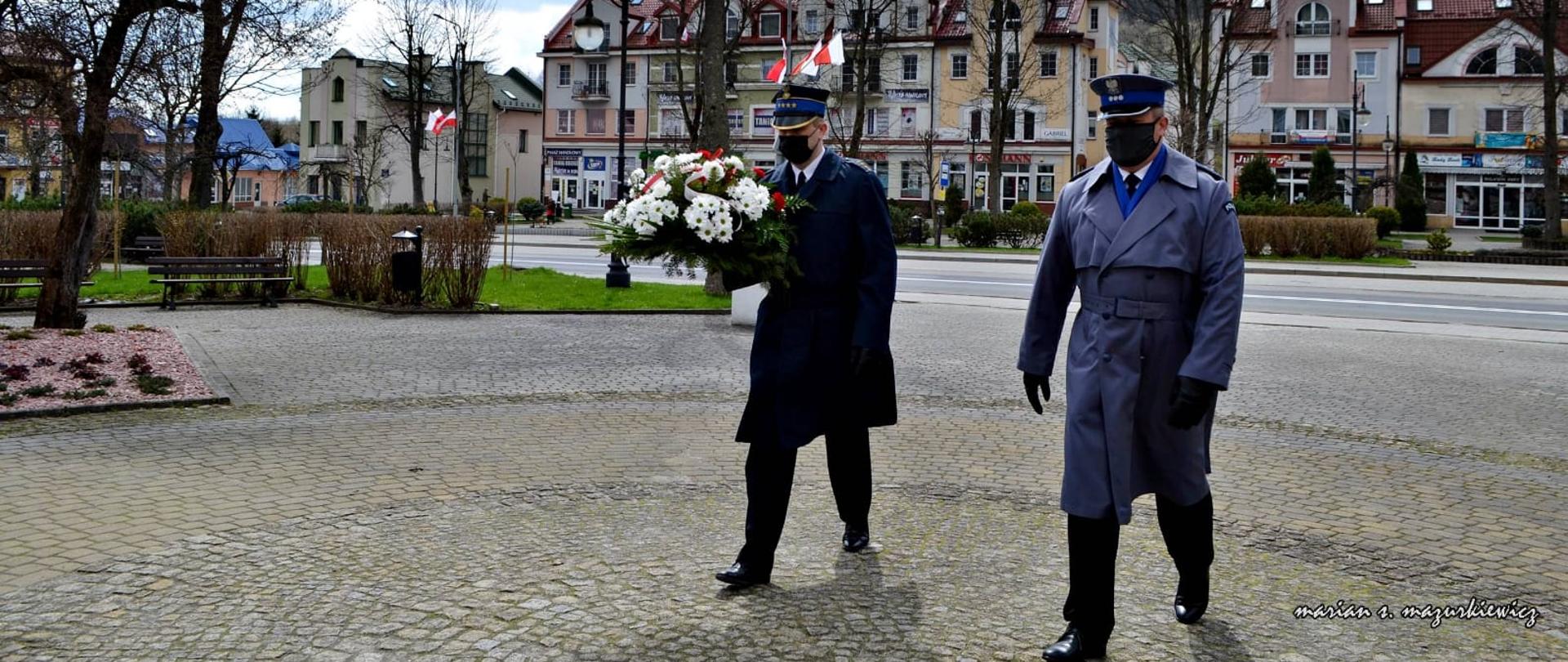 Na zdjęciu widać Komendantów Powiatowych Państwowej Straży Pożarnej oraz Policji w Ustrzykach Dolnych w mundurach galowych którzy idą w kierunku pomnika. Komendant Powiatowy PSP niesie w ręku kwiaty. W tle widać rynek, budynki oraz powiewające na wietrze flagi. 