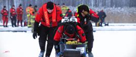 6. Strażacy w kombinezonach lodowych stoją przy saniach ratowniczych ze sprzętem technicznym i linami, koordynując działania na zamarzniętym jeziorze.
