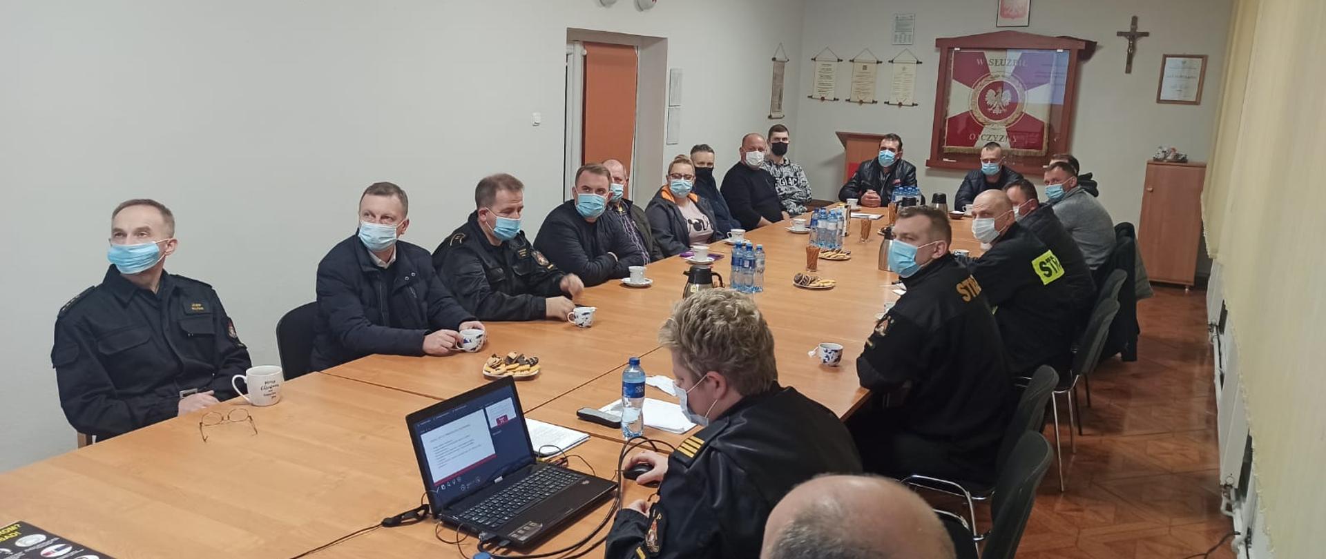 Zdjęcie przedstawia grupę ludzi siedzących w świetlicy przy stołach w maseczkach, którzy słuchają prowadzącego spotkanie. W tle widzimy na ścianie godło Polski, sztandar KP PSP Świdwin oraz krzyż.