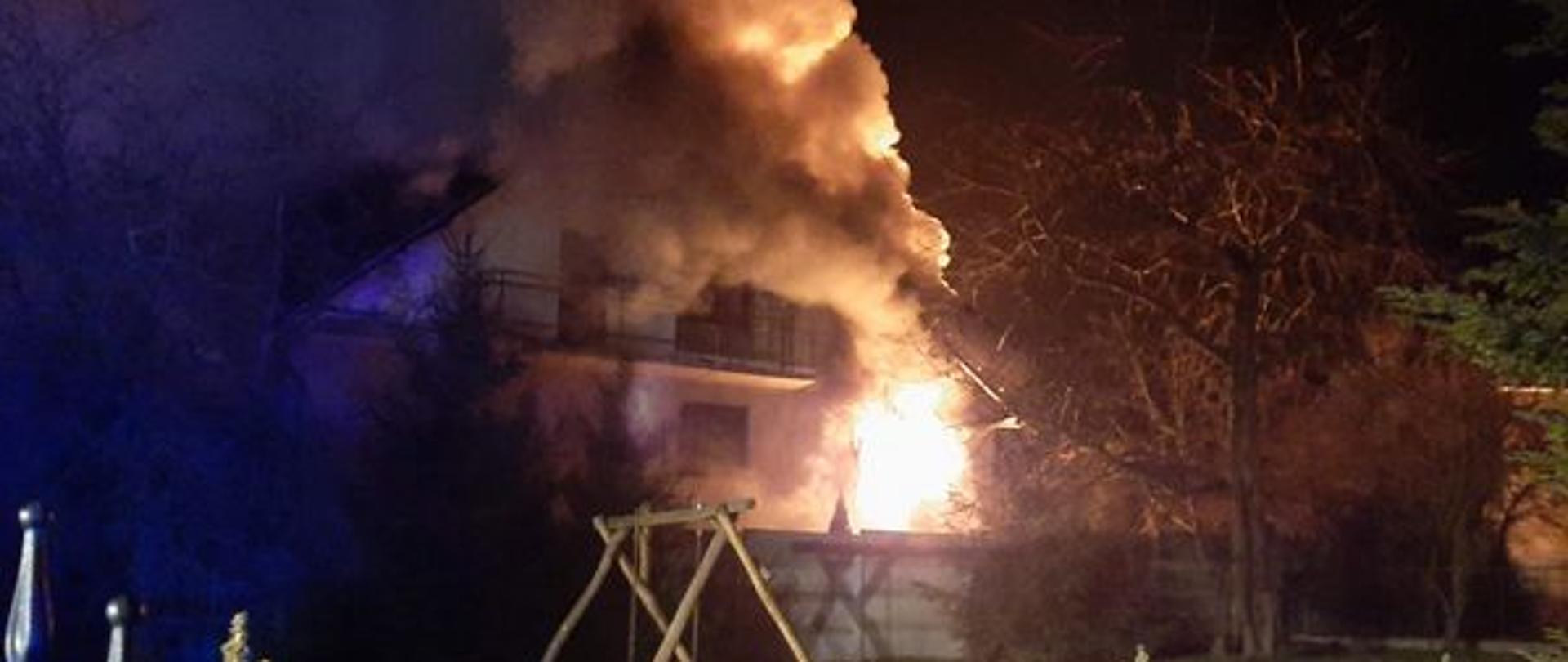 Dom dwukondygnacyjny częściowo objęty pożarem. Widoczne kłęby dymu. Pora wieczorna.