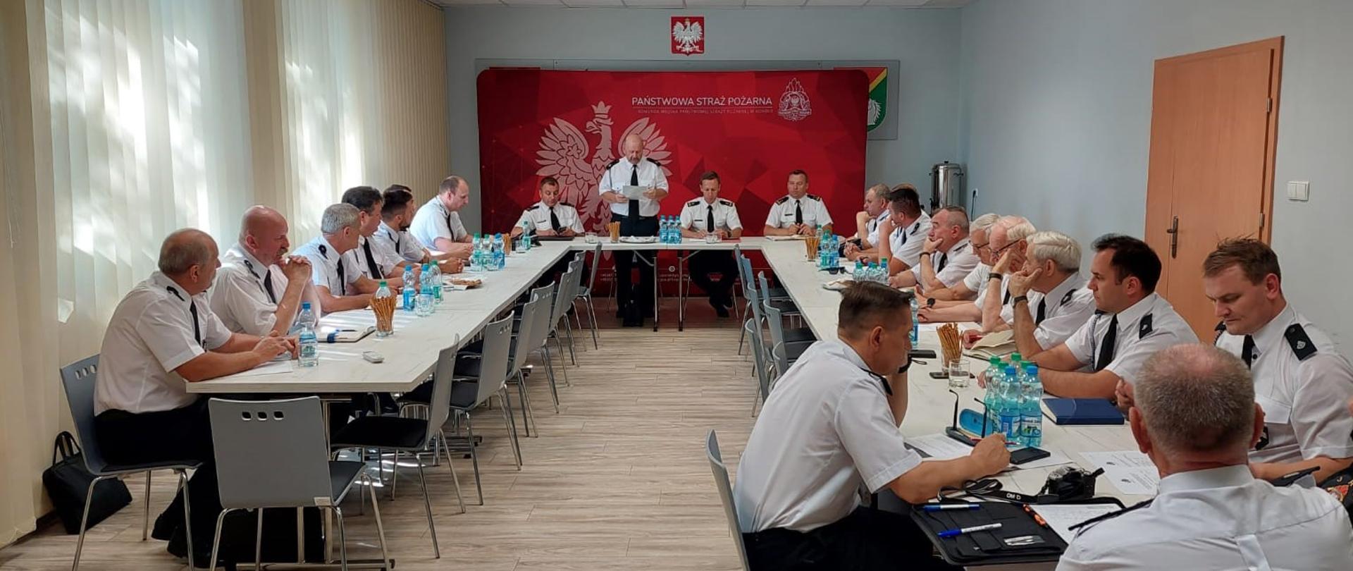 Zdjęcie przedstawia druhów OSP siedzących przy stole, ustawionym w kształcie litery U. Przy środkowym stole siedzi Zastępca Wielkopolskiego Komendanta Wojewódzkiego PSP, Prezes Powiatowy ZOSP RP, Komendant Miejski PSP i Zastępca Komendanta Miejskiego PSP. W tle ścianka komendy z godłem.