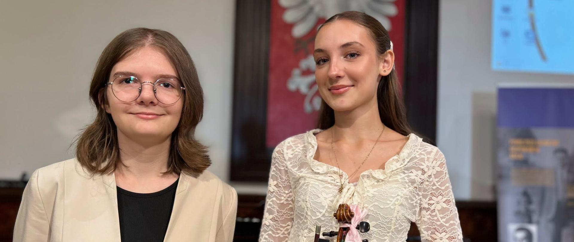 Dwie uczennice — Daria Kopacz i Julia Kurnal trzymająca skrzypce — pozują do zdjęcia w Sali Królewskiej po występie podczas memoriału „Mieleccy Sprawiedliwi wśród narodów świata", w tle herb Polski.
