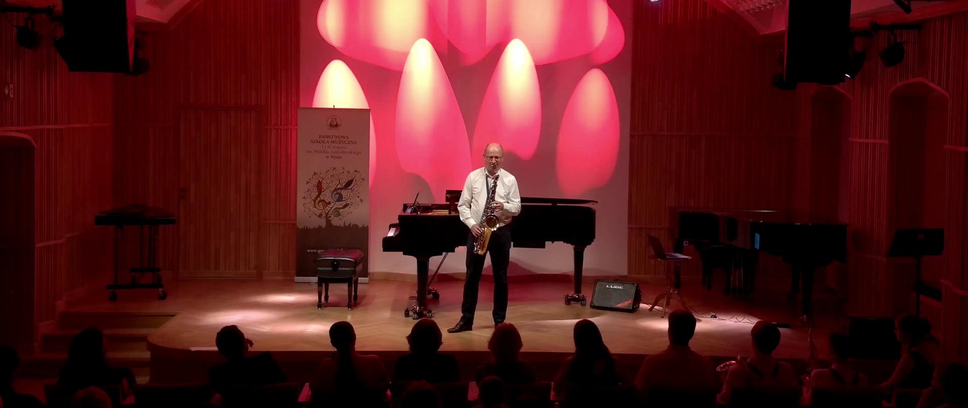Zdjęcie. Na estradzie sali koncertowej występuje solo saksofonista. Przed nim widać publiczność. w tle stoi fortepian, a na ścianie wyświetlona jest grafika w kolorze czerwonym.