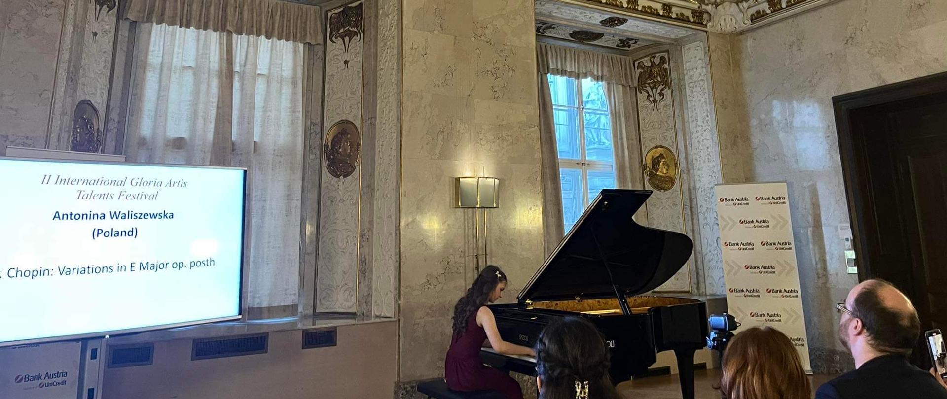 Pianistka siedzi przy fortepianie w sali Ratusza Miejskiego w Wiedniu ubrana w czerwoną wieczorową suknię., po lewo ekran, na którym wyświetlone jest jej nazwisko i program 
