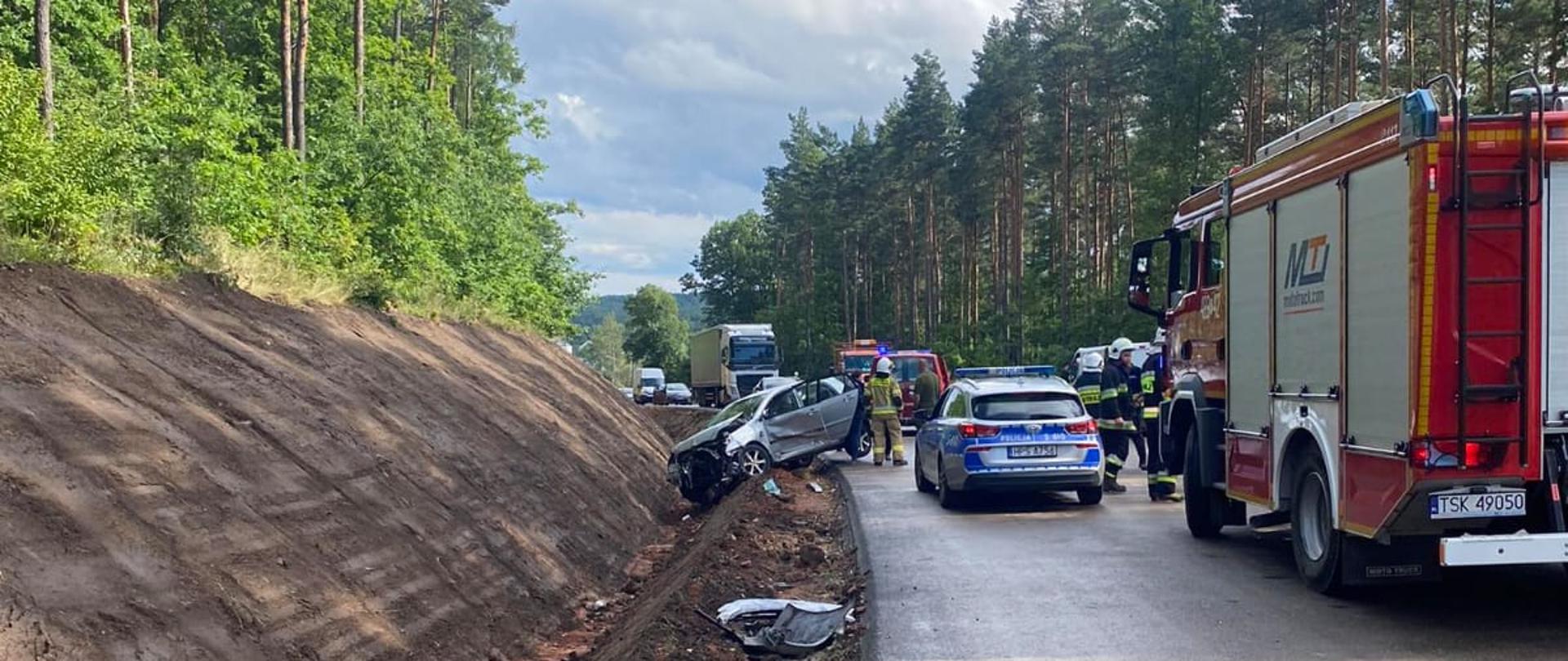 Zdjęcie przedstawia samochód osobowy po wypadku samochodowym, który znajduje się w rowie. Obok niego stoją strażacy OSP i radiowóz Policji oraz samochód pożarniczy. W tle widać inne pojazdy.
