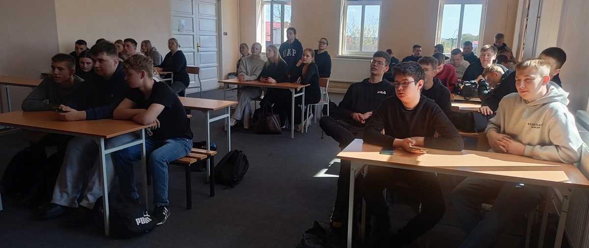 Zdjęcie przedstawia uczniów zgromadzonych w sali lekcyjnej podczas trwania konkursu. Uczestnicy siedzą w ławkach, skupieni i zaangażowani, wielu z nich korzysta z telefonów, odpowiadając na pytania quizu. W sali panuje spokojna atmosfera sprzyjająca rywalizacji i wspólnej zabawie.